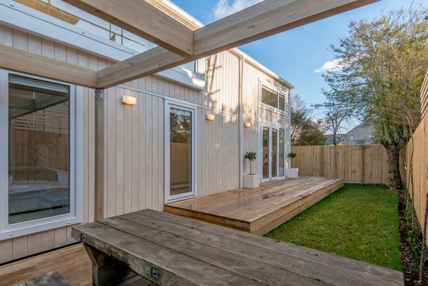 A bright exterior courtyard view of a modern timber-clad home, featuring a raised timber deck, pergola beams, large glazed doors and windows, and a fenced garden courtyard. The design has clean vertical weatherboard cladding, simple detailing, and strong indoor-outdoor connections.