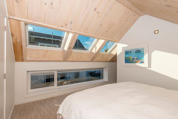 Bright attic-style bedroom with a large bed, pale carpet, white walls, and a distinctive sloped timber-lined ceiling incorporating multiple skylights and roof windows. Horizontal windows beneath the eaves provide additional daylight and views of neighbouring buildings. The room has a simple, airy contemporary feel with a framed artwork on the wall.