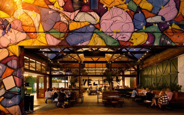 A vibrant commercial dining and hospitality interior viewed through a large abstract stained-glass-style mural or illuminated graphic facade. The space has exposed dark steel framing, timber flooring and furnishings, extensive glazing, warm pendant and string lighting, communal tables, banquette seating, and multiple diners. A large central sign is visible in the background, while colourful organic geometric artwork dominates the upper and side portions of the image.