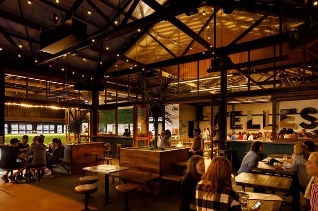 A warmly lit contemporary industrial-style restaurant or hospitality interior with open-plan dining, timber booth seating, bar-height tables, and numerous patrons. Black exposed steel roof trusses, suspended pendant and track lighting, timber wall and ceiling elements, and extensive internal glazing create a dramatic, layered atmosphere. Large graphic wall lettering and indoor plants add visual interest.