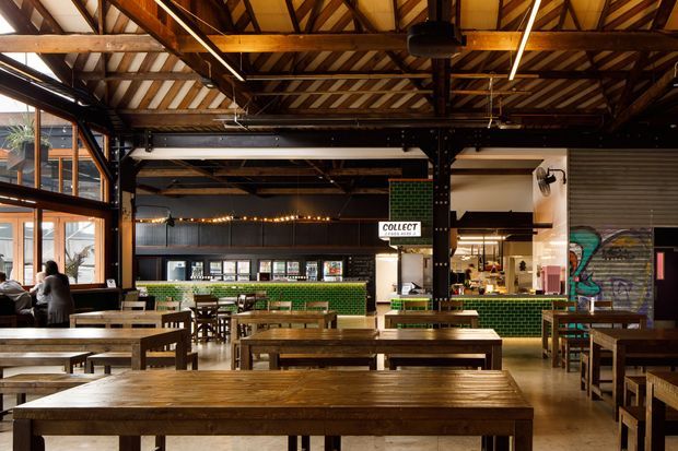 A spacious industrial-style commercial dining hall or food venue with long communal timber tables and benches, exposed timber roof trusses, black steel structural columns, polished concrete flooring, large steel-framed windows, and a green glazed-tile service counter. A visible 'COLLECT' sign and food-service areas reinforce the hospitality setting, while a roller door and colourful mural add warehouse character.