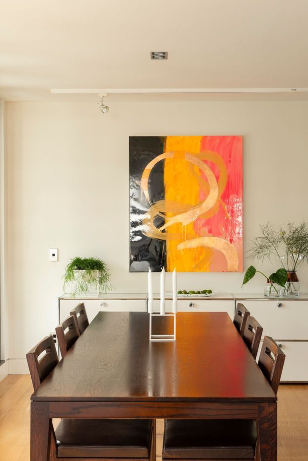 A warm, refined dining room viewed toward a long dark timber dining table with matching chairs. A pale neutral wall features a large abstract artwork in orange, red, black and cream, while a low built-in sideboard displays candles, plants and decorative objects. The room has a simple painted ceiling with track lighting and light timber flooring.