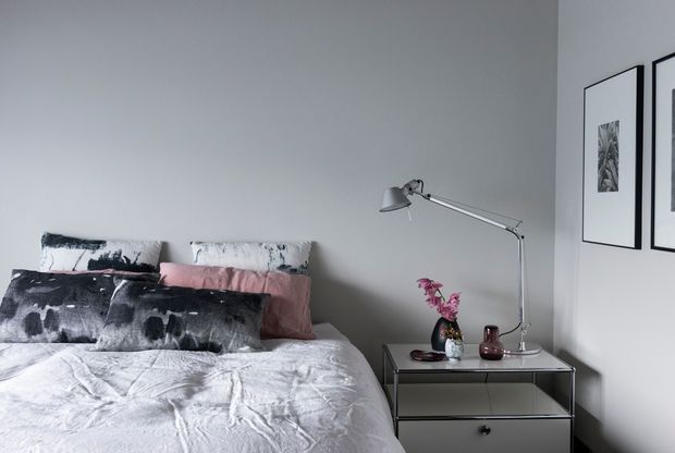 A styled bedroom with a low-profile bed dressed in layered white, grey, black, and blush bedding. A grey bedside table with open shelving and a drawer supports a slim articulated metal lamp, small vases, and flowers. Pale grey walls and black-framed monochrome artwork create a calm contemporary setting.