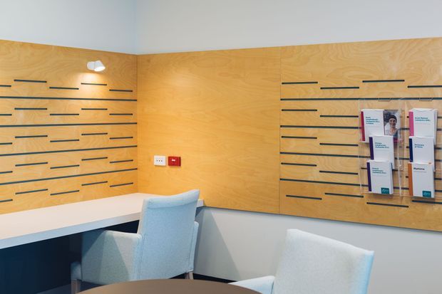 A bright contemporary reception or consultation area with warm timber slat-wall panels, integrated dark horizontal display rails, a wall-mounted brochure holder, and a built-in white desk or counter. Pale upholstered chairs sit in front of the counter, with a small wall light and minimal electrical controls adding functional detail.