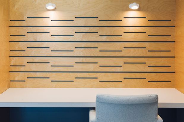 Compact built-in workspace with a warm plywood feature wall, horizontal dark recessed handles or trim lines, integrated round downlights, and a white desk surface. A light upholstered chair faces the desk, creating a clean, tailored home office or study setting.
