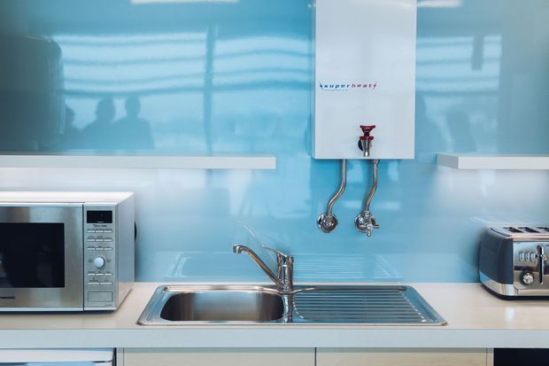 Compact modern kitchen or kitchenette with a glossy turquoise glass splashback, pale benchtops, stainless-steel sink and drainer, wall-mounted hot-water unit, microwave, and toaster. The space is brightly lit with clean, streamlined finishes.