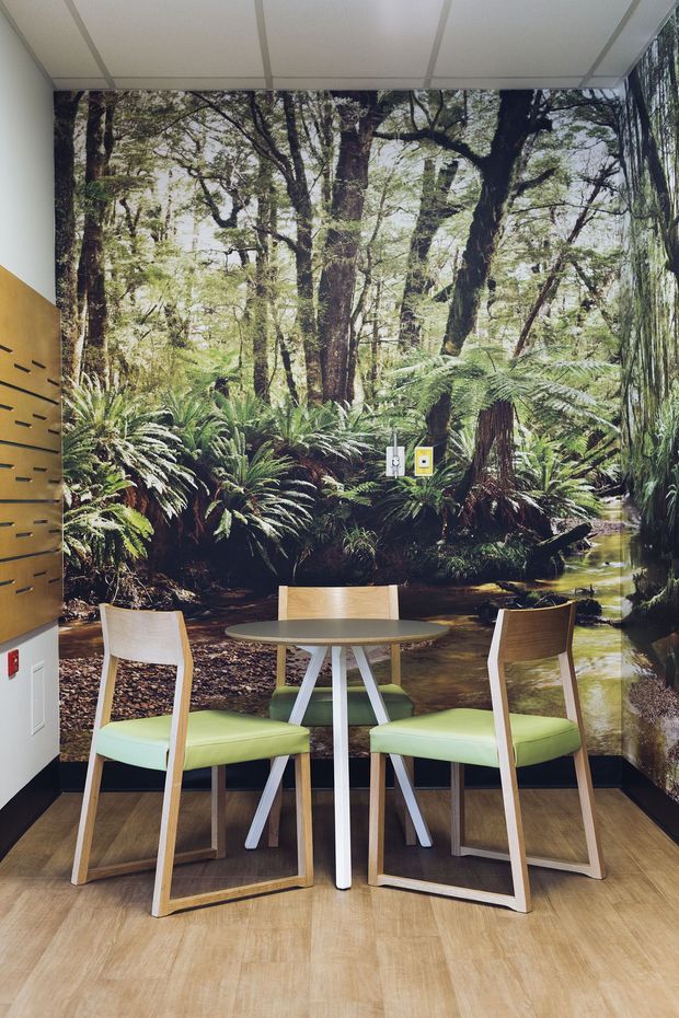A compact contemporary interior dining nook with a small round table and four light timber-framed chairs upholstered in pale green. A large forest and fern mural spans the rear and side walls, creating a strong biophilic focal point. The space has warm wood-look flooring, white acoustic ceiling panels, a mustard-yellow built-in wall section with horizontal black pulls, and a narrow black skirting detail.