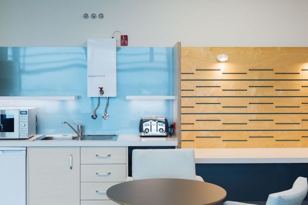Compact contemporary kitchenette and dining nook with pale cabinetry, stainless-steel sink and tapware, a vivid blue glass splashback, and a warm timber feature wall with horizontal dark inlays. A small round dark dining table and upholstered chairs occupy the foreground.