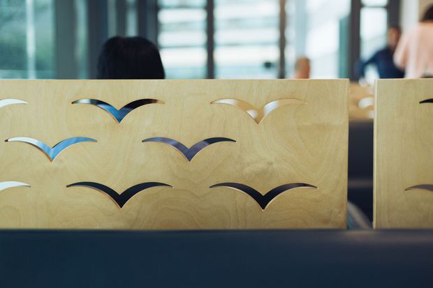 Close-up view of a light timber reception or privacy screen featuring repeated stylised bird-shaped cut-outs. The screen sits in a contemporary commercial interior, with blurred glazing and people in the background and a dark blue upholstered or solid foreground element.