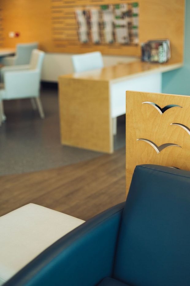 Interior view of a contemporary reception or waiting area, featuring custom light-wood joinery and screening panels with curved metal inlays, teal upholstered seating in the foreground, a pale reception desk, white lounge chairs, warm timber wall cladding, and dark flooring.
