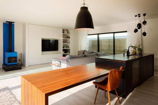 Open-plan contemporary kitchen, dining and living area with white built-in cabinetry, a large warm timber dining table, dark kitchen island, grey sectional sofa, black pendant lighting and expansive sliding glass doors. A vivid blue built-in fireplace feature and orange upholstered dining chair add strong colour accents.