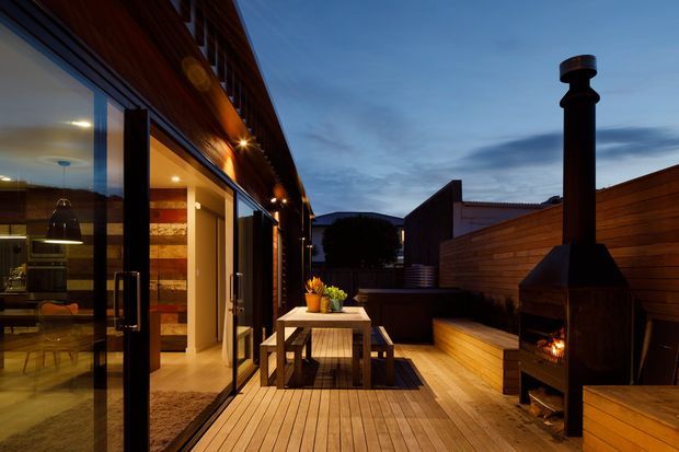 Twilight view of a contemporary timber-clad outdoor deck adjoining a glazed interior. The deck features a dining table, built-in timber bench seating, planter boxes, warm exterior lighting, and a freestanding outdoor fireplace or chiminea. Floor-to-ceiling sliding glass doors reveal a dining area inside, while a timber privacy screen encloses the terrace.