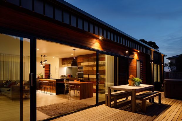 Twilight view of a contemporary single-storey home with a long dark metal-clad roofline and warm timber-clad facade. Floor-to-ceiling sliding glass doors open onto a timber deck with a built-in picnic-style outdoor dining table. The illuminated interior visibly contains an open-plan kitchen and living area, with timber cabinetry, a timber feature wall, pendant lighting and a dining table.