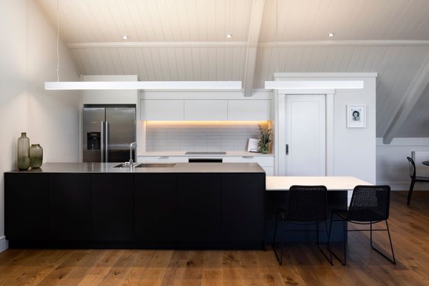 A bright, contemporary open-plan kitchen and dining area beneath a sloped white paneled ceiling. The kitchen features a large matte-black island with integrated sink, stainless-steel refrigerator, white cabinetry, a warm illuminated tiled splashback, and timber flooring. A light dining table with black wire-frame chairs sits beside the island, with a white paneled door and built-in storage visible in the background.