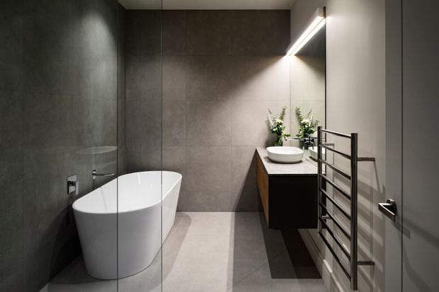 Contemporary bathroom with a freestanding white bathtub, large-format grey wall and floor tiles, frameless glass shower screen, floating dark wood vanity with a white vessel basin, and a wall-mounted heated towel rail. A large mirror with integrated linear lighting and small plants adds a refined, spa-like feel.