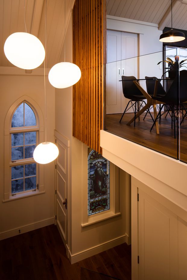 A tall, warm-toned interior stair hall with a double-height void and mezzanine dining area. The space features dark timber flooring, white painted wall panelling, a vertical timber slat screen, black-framed glass balustrade, sculptural pendant lights, an arched multi-pane window, and a decorative stained-glass window. A compact dining setting is visible on the upper level.