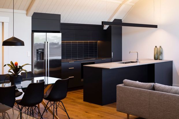 Open-plan kitchen and dining area with a large black island, dark flat-panel cabinetry, stainless-steel refrigerator, tiled splashback, and integrated sink. The space includes a timber floor, white pitched-panel ceiling, pendant lighting, dining table with black chairs, and an adjacent grey sofa, creating a contemporary, high-contrast interior.