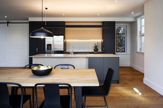 A bright, contemporary open-plan kitchen and dining area. The kitchen features dark flat-panel cabinetry, a light timber-toned benchtop and splashback, integrated appliances, a central island, and white horizontal tile detailing. A timber dining table with black chairs occupies the foreground. Timber flooring, a black linear pendant light, recessed ceiling lights, a sliding barn-style door, and a stained-glass feature window add visual interest.