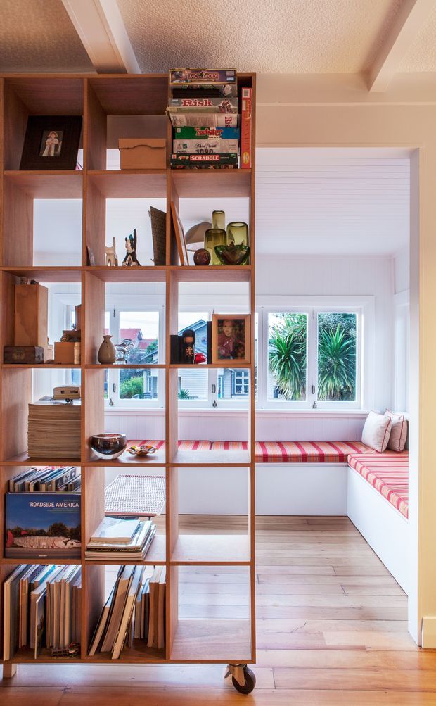 Bright interior view of a compact window-lined reading nook or sunroom, partially screened by a tall open timber shelving unit on casters. The shelving displays books, board games, photographs, ceramics, and decorative objects. Built-in bench seating wraps beneath the windows and is upholstered in vivid red and pink striped fabric with cushions. Pale timber flooring, white walls, white-framed windows, and a textured white ceiling create an airy residential space.