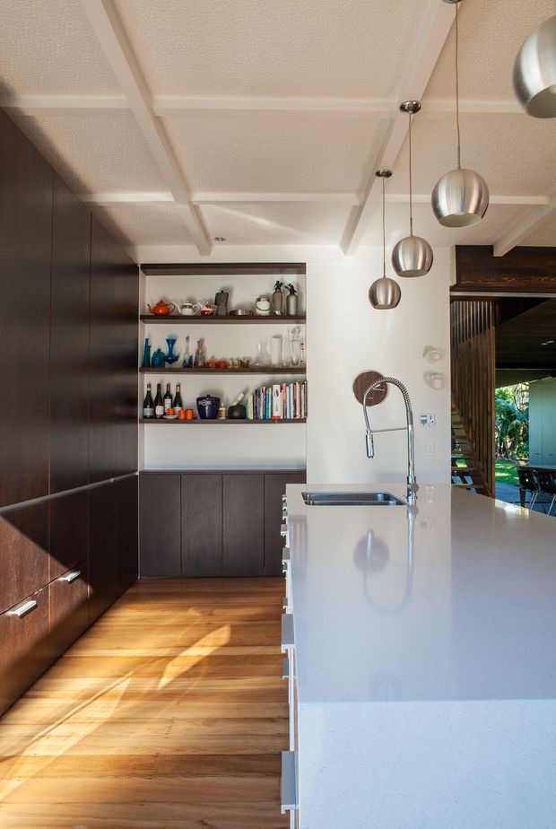 A bright contemporary kitchen with a long white island and integrated sink, dark timber cabinetry, open shelving displaying glassware and kitchen items, and warm timber flooring. Three spherical metallic pendant lights hang above the island. The textured white coffered ceiling and open connection to an outdoor area are prominent.