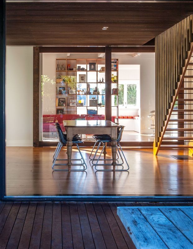 A warmly lit contemporary dining area viewed from an exterior timber deck, with a rectangular dining table and chairs in the foreground. Beyond it is a glass-framed living space with a red sofa, open shelving, and large windows. A prominent timber staircase with vertical balustrade rises along the right side, while dark timber ceiling boards, exposed structural posts, and polished timber flooring create a cohesive material palette.