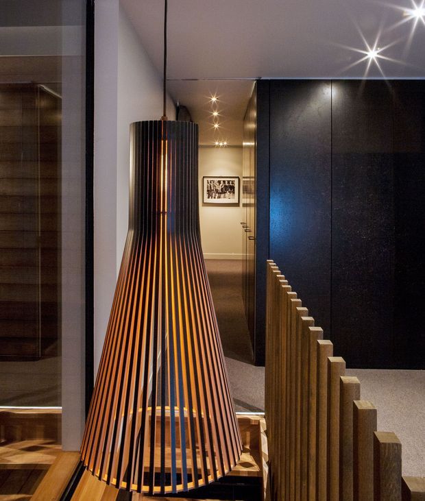 Interior architectural photograph focused on a striking sculptural pendant light made from evenly spaced dark timber slats with a warm amber glow. It hangs in a refined hallway or open-plan interior, with dark textured wall panelling and cabinetry on the right, timber flooring and joinery, carpeted circulation space, recessed spotlights, and a framed artwork in the background. A low timber slat screen or balustrade runs along the right side, creating layered depth and strong vertical rhythm.