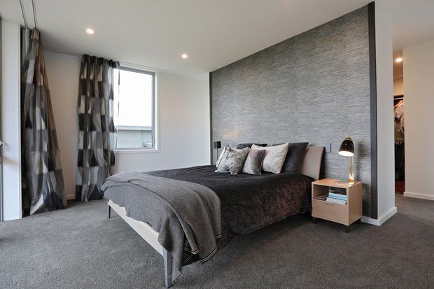 A contemporary bedroom with a low platform bed dressed in layered charcoal and grey bedding, patterned cushions, and a textured grey feature wall behind the bed. Floor-to-ceiling glazing and a side window bring in natural light, while patterned curtains, recessed ceiling lights, a compact timber bedside table, and a black table lamp complete the room. An open walk-in wardrobe is visible at the right.
