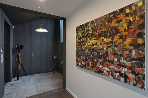 A contemporary entry hall viewed from the adjoining interior, with dark vertical wall panelling, large-format pale stone-look floor tiles, a sculptural pendant light, and a large colourful abstract artwork mounted on a light neutral wall. A narrow doorway and decorative objects add depth to the compact foyer.