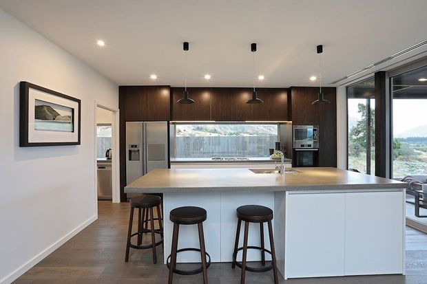 Contemporary open-plan kitchen with a large pale island and three dark timber bar stools, dark wood full-height cabinetry, integrated stainless-steel appliances, and a wide horizontal window above the benchtop. Recessed and pendant lighting illuminate the clean, neutral space, while full-height glazing on the right opens views toward the landscape and outdoor seating.