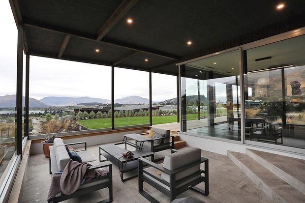 A covered outdoor lounge or terrace with a dark exposed-beam ceiling, recessed lighting, expansive black-framed glazing, and contemporary outdoor seating arranged around low tables. The terrace overlooks a landscaped lawn and distant mountainous scenery, with broad steps leading to a glazed interior space.
