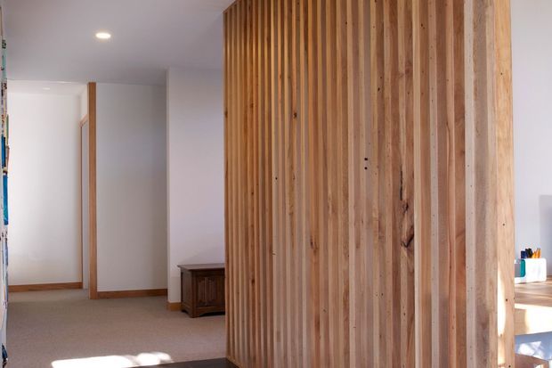 Interior view of a bright, contemporary home featuring a prominent vertical timber slat partition or feature wall. The space includes white painted walls and ceiling, warm timber trims and joinery, carpeted flooring, recessed lighting, and a partial glimpse of a desk or workspace at right.