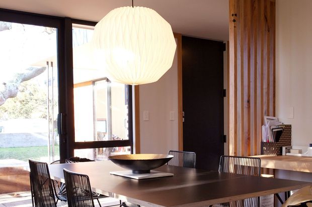 A contemporary dining area with a large dark rectangular dining table, slim black wire-frame chairs, and an oversized white pendant light. Floor-to-ceiling glazing opens toward an outdoor area, while a vertical timber slat feature wall and built-in wood-toned cabinetry add warmth. The palette is neutral with dark timber, white walls, and abundant natural light.