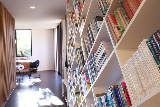 A bright, narrow interior corridor lined with extensive white built-in bookshelves filled with books. A compact desk and chair sit near a large window at the far end, while vertical timber slats define the left wall. The space has a dark timber floor, white ceiling, recessed lighting, and a calm contemporary study or library character.