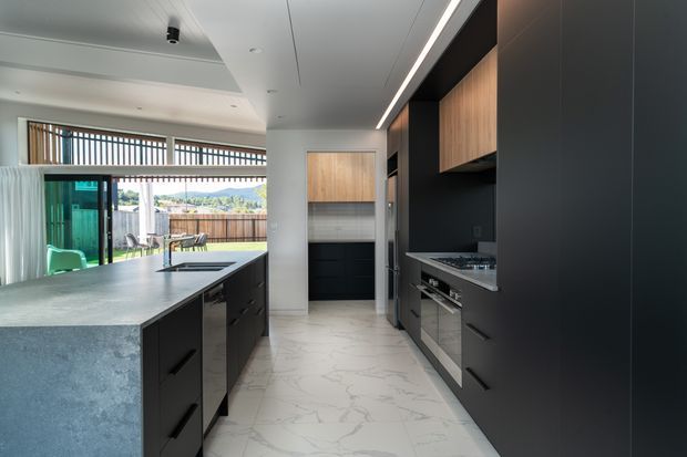 Contemporary galley-style kitchen with a large grey island, dark matte cabinetry, warm timber overhead cupboards, integrated stainless-steel appliances, and a white marble-look tiled floor. Full-height glazing opens the space toward an outdoor area, while linear ceiling lighting and clean built-in joinery create a refined modern interior.