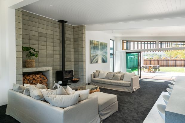 Bright contemporary living room with a large white sectional sofa, secondary seating, dark carpet, and a prominent grey blockwork fireplace with integrated timber storage and black flue. The open-plan space connects directly to a covered outdoor entertaining area through wide glazed doors, with a dining setting visible outside. White-lined ceilings, pale walls, and abundant daylight create an airy, comfortable interior.