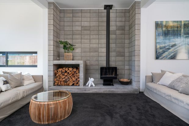 A bright contemporary living room centred on a freestanding black wood-burning stove with a tall flue, set against a full-height grey tiled fireplace wall and concrete hearth. Built-in log storage sits to one side, with pale sofas, a rounded slatted timber coffee table, dark carpet, and a framed abstract artwork.
