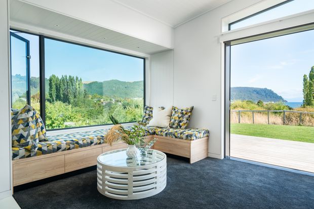 Bright, contemporary sunroom or living area with a built-in timber window seat and matching low lounge bench upholstered in a bold geometric blue, mustard and grey pattern. A round white slatted coffee table, cushions and plant sit on a dark carpet. Large corner glazing and a wide glazed door open to a timber deck and landscaped lawn, with green hills and water visible beyond.
