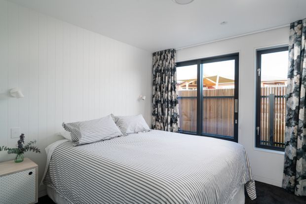 Bright, compact bedroom with a neatly made bed dressed in fine grey-and-white striped bedding, white vertical wall panelling, dark carpet, and large black-framed windows and sliding doors opening to a fenced outdoor area. Patterned floor-length curtains add contrast, while a small bedside table and plant complete the room.