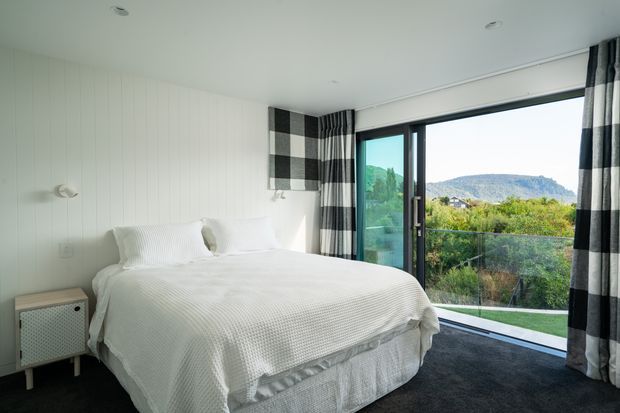 Bright contemporary bedroom with a low-profile bed dressed in white textured bedding, white vertical wall panelling, and bold black-and-white checked curtains. Full-height sliding glass doors open to a balcony or terrace with views toward a green, hilly landscape. A small light-toned bedside cabinet and discreet wall lights complete the room.