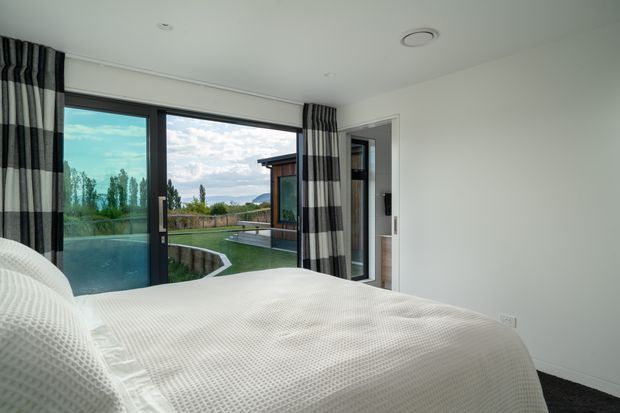 Contemporary bedroom viewed from beside the bed, with a white textured bedspread, pale walls, dark carpet, and full-height glazed sliding doors opening to a landscaped outdoor area with a pool. Bold black, white and grey striped curtains frame the glazing, and a secondary glass door is visible to the right.