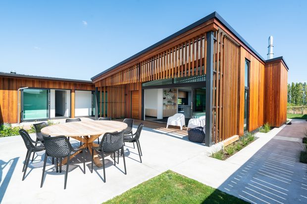 Bright exterior view of a contemporary single-storey home arranged around an outdoor entertaining area. The building features warm vertical timber screening and cladding, large glazed openings, a low-pitched dark roof, and a paved terrace with outdoor dining furniture. A covered open-plan area appears to connect to the interior.