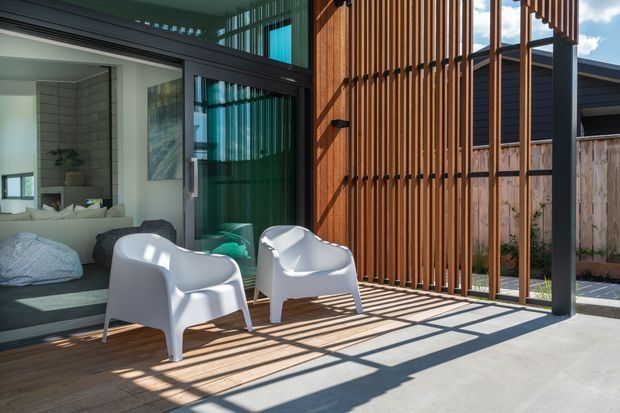 A sunlit contemporary outdoor terrace or patio beside a glazed living area and bedroom. The space features a concrete floor, timber decking, a vertical timber batten privacy screen, dark metal framing, full-height glazing, and two white modern lounge chairs. A bedroom and living area are visible through the open glazing.