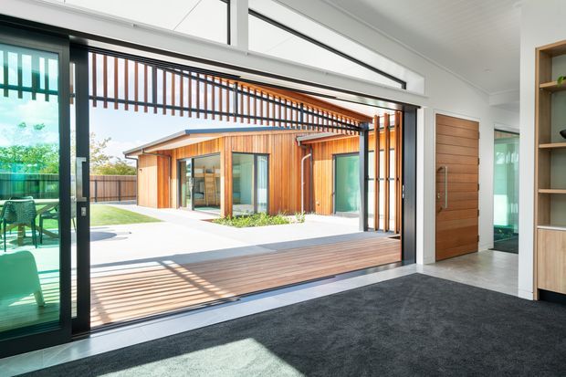 Bright contemporary interior opening onto a landscaped courtyard through expansive black-framed glazed doors. The room has dark carpet, white walls and ceiling, a timber door, and built-in timber shelving. Outside, a timber-clad pavilion with a low pitched roof, covered timber deck, and strong vertical timber screening is visible. The open glazing creates a seamless indoor-outdoor connection with abundant natural light.