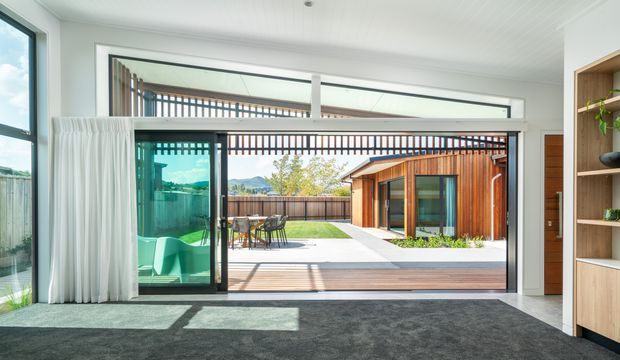 Bright contemporary interior opening through full-height sliding glass doors to a timber-decked courtyard and lawn. The room has a dark carpeted floor, white walls and ceiling, sheer curtains, built-in light timber shelving, and strong indoor-outdoor connection. Outside is a compact timber-clad pavilion with a glazed door, covered dining area, and linear slatted screening.