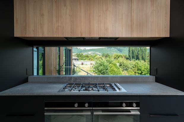 Contemporary kitchen with dark matte cabinetry and a broad grey stone benchtop, centred on a gas cooktop and twin built-in ovens. A full-width picture window behind the cooktop frames a leafy hillside view, while a large light timber overhead cabinet adds warmth and contrast.