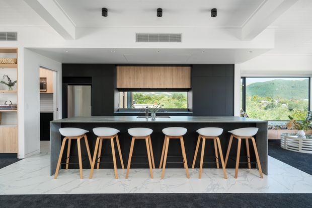 Contemporary open-plan kitchen dominated by a long black island with six timber-legged bar stools. The kitchen features dark matte cabinetry, a timber roller blind and warm timber shelving, a pale veined stone-look floor, integrated appliances, and expansive windows overlooking a green landscape. White ceiling beams and recessed lighting create a bright, spacious interior.