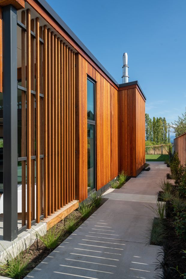 A contemporary building exterior viewed along a landscaped concrete walkway. The facade combines rich vertical timber cladding and closely spaced timber screening with dark metal framing, tall narrow glazing, and a low-pitched dark roof. Ornamental grasses and planting border the path, while a metal flue projects from the roof against a clear blue sky.