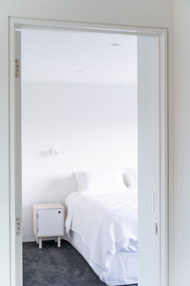 A bright, minimalist view through a doorway into a white bedroom. The room contains a white bed with pillows and textured bedding, a small light-coloured bedside cabinet, dark grey carpet, white walls, recessed ceiling lights, and white-painted door and wall trim.