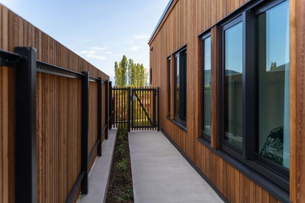 A narrow, enclosed side passage between a timber-clad building and a matching timber privacy fence. The passage has a smooth concrete walkway, a slim planted garden strip, black-framed windows and glazing, and a timber gate at the far end. The exterior is contemporary and warm, with strong linear detailing and clear blue-sky daylight.
