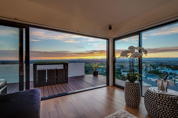 A contemporary living room with expansive corner glazing opening onto a timber-clad balcony or terrace. The room overlooks a coastal or waterfront urban landscape at sunset, with a small outdoor dining setting beyond the doors. Inside are timber-look flooring, neutral walls, a dark upholstered sofa, and sculptural textured planters with orchids. The image has a refined, warm atmosphere and emphasizes the panoramic outlook and indoor–outdoor connection.